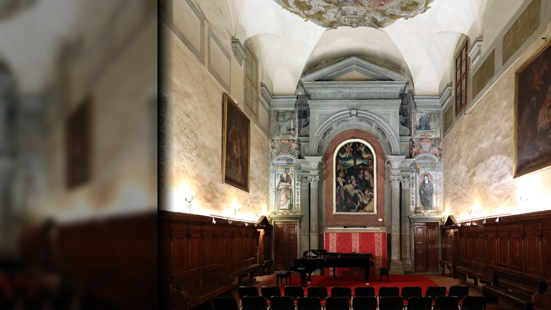 The nave of Santa Monaca church in Florence set for a concert: rows of chairs and a grand piano on a red carpet before a stone-framed altar with a painted altarpiece, wooden choir stalls along both walls and a frescoed vault above